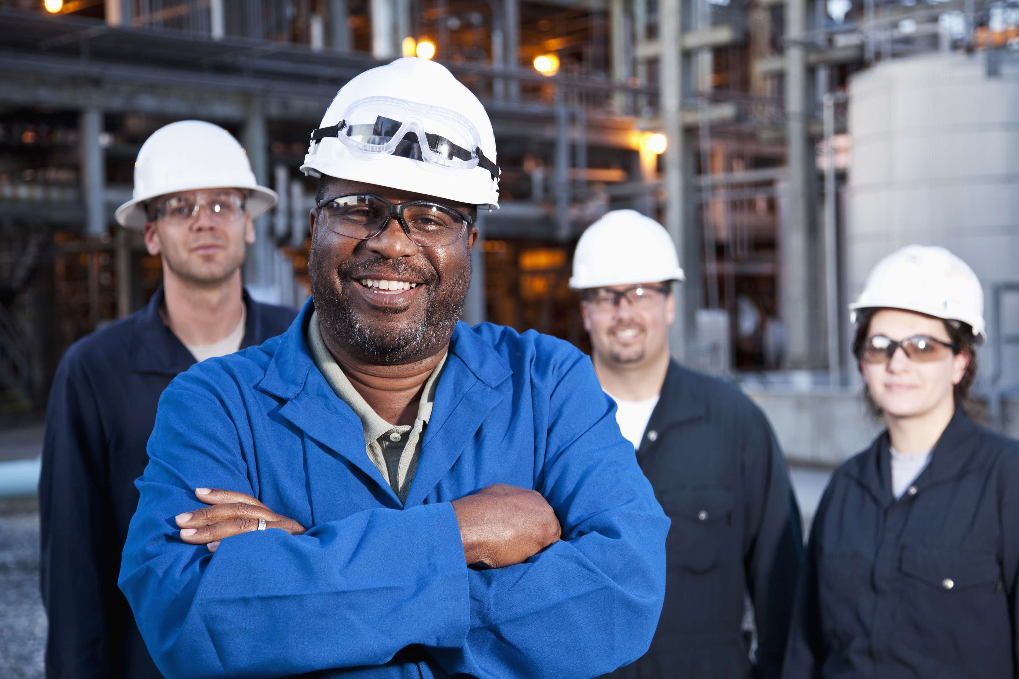 A group of people, one in the foreground, three in the background, wearing goggles and safety helmets, stand in front of pipes and a storage tank.