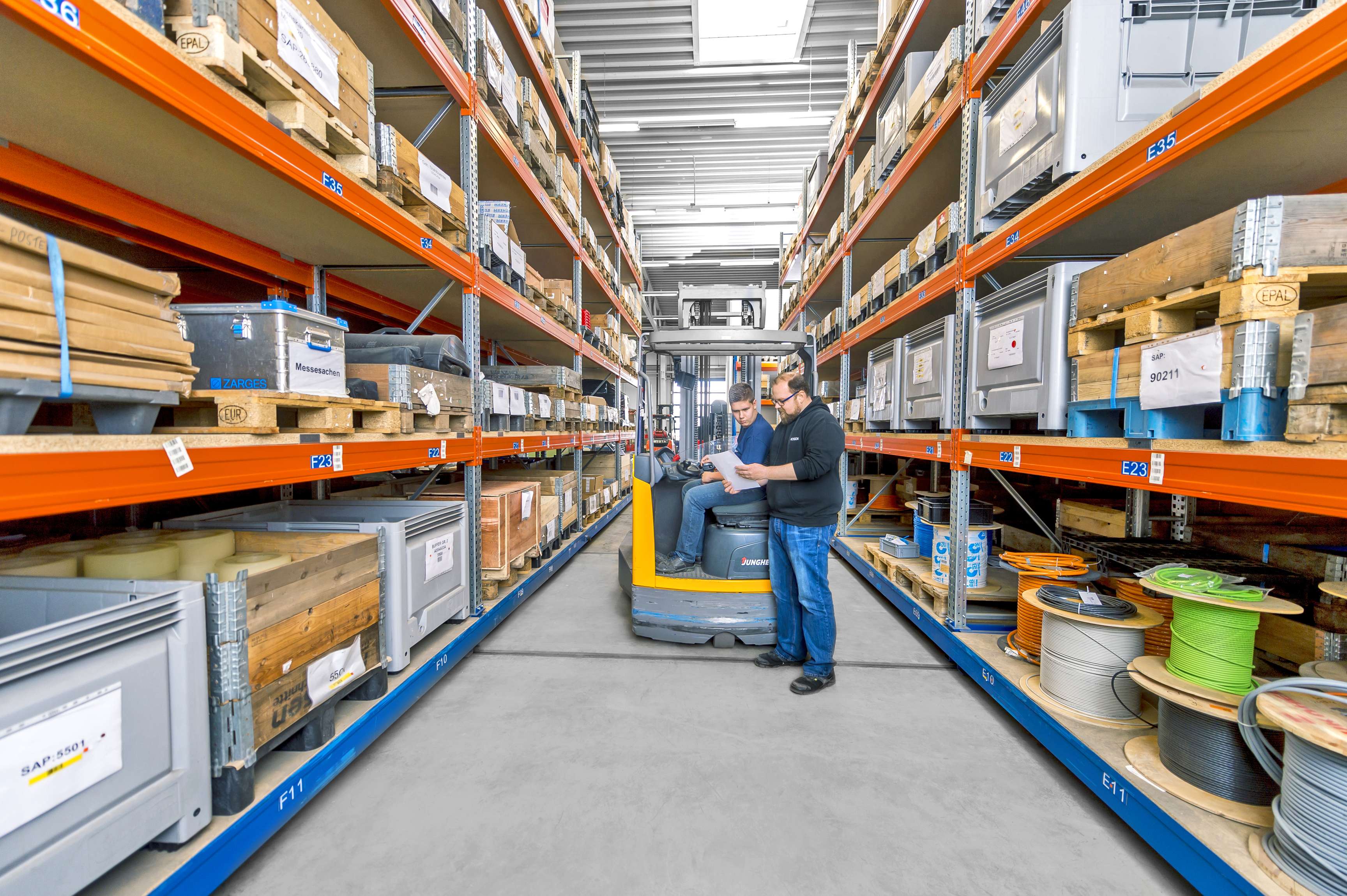 Zwei Mitarbeiter stehen in der Mitte von Hochregalen, in denen Kartons und Pakete stehen.