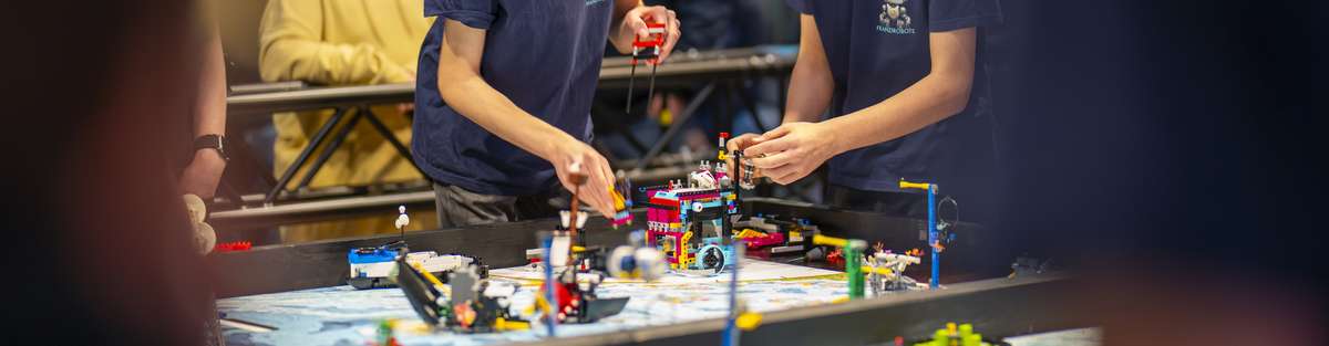 Two boys are working on their LEGO robot at a table. Onlookers are watching them.