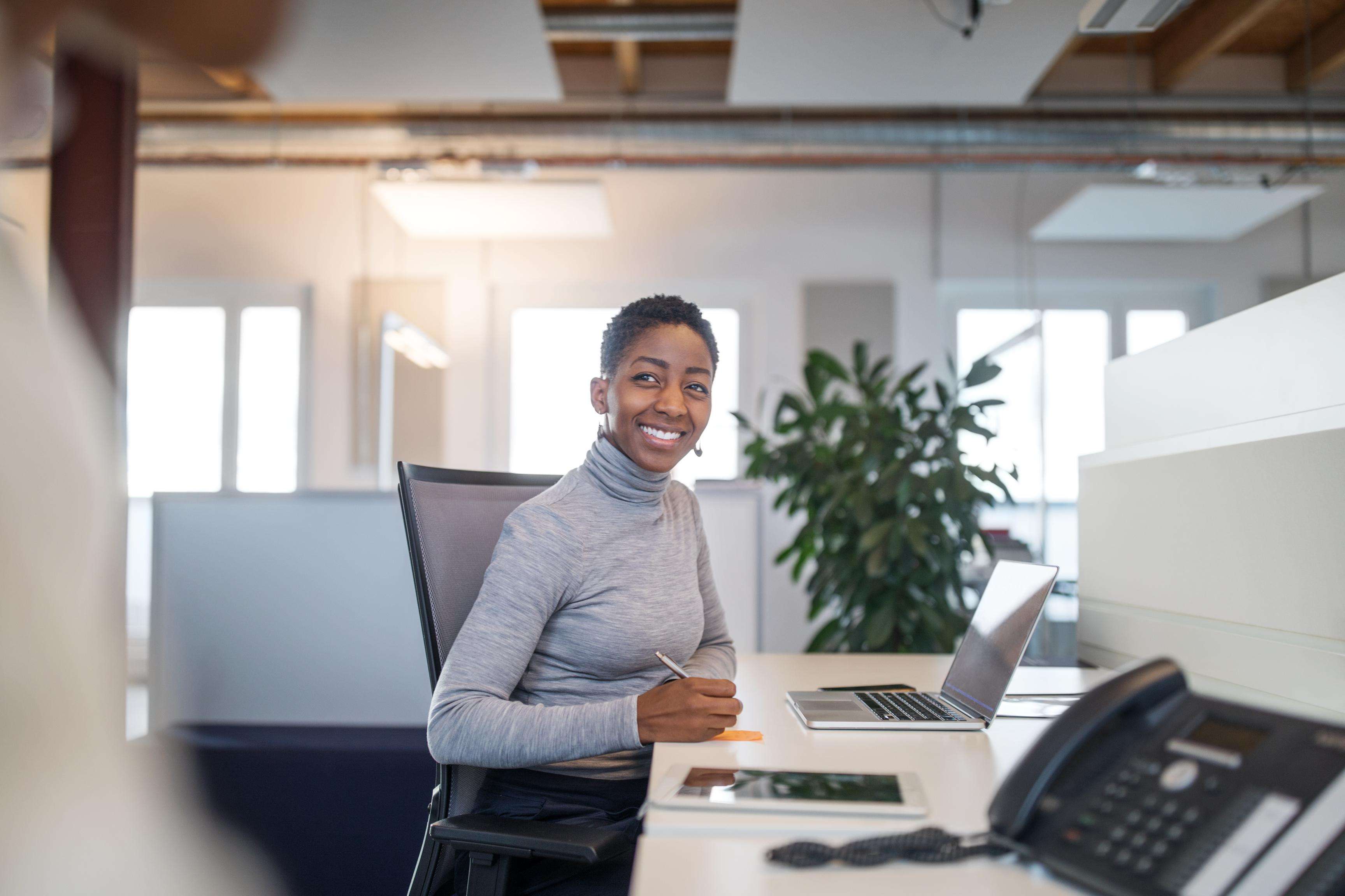 Woman sitting at a desk in front of a laptop while taking notes with a pen and smiling sideways at the camera.