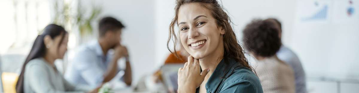 Woman sitting in the office in front of her laptop, taking notes and smiling at the camera, while her colleagues can be seen in the background.