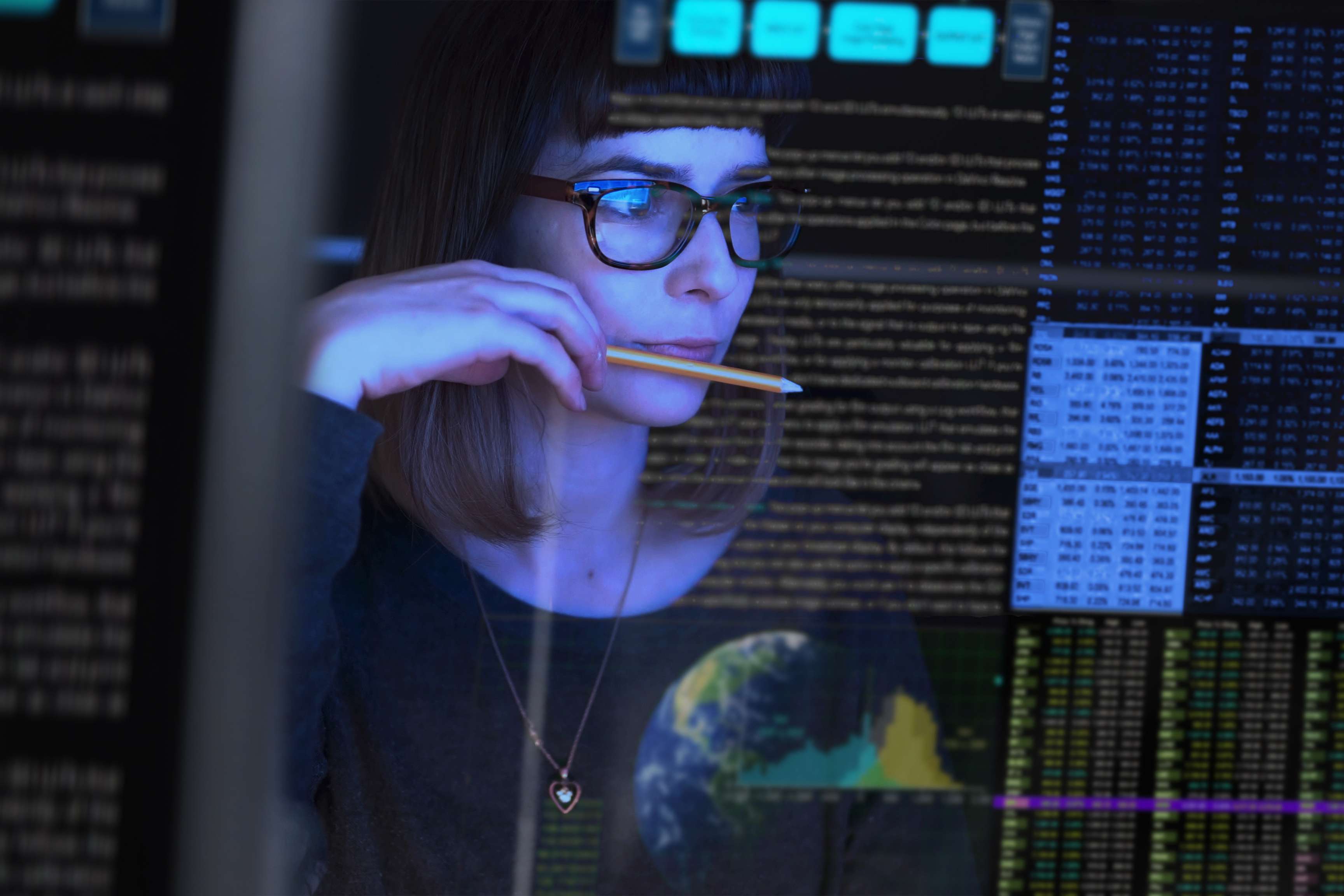 Woman wearing glasses and holding a pencil in her hand and putting it to her lips while looking at a screen with data sets.