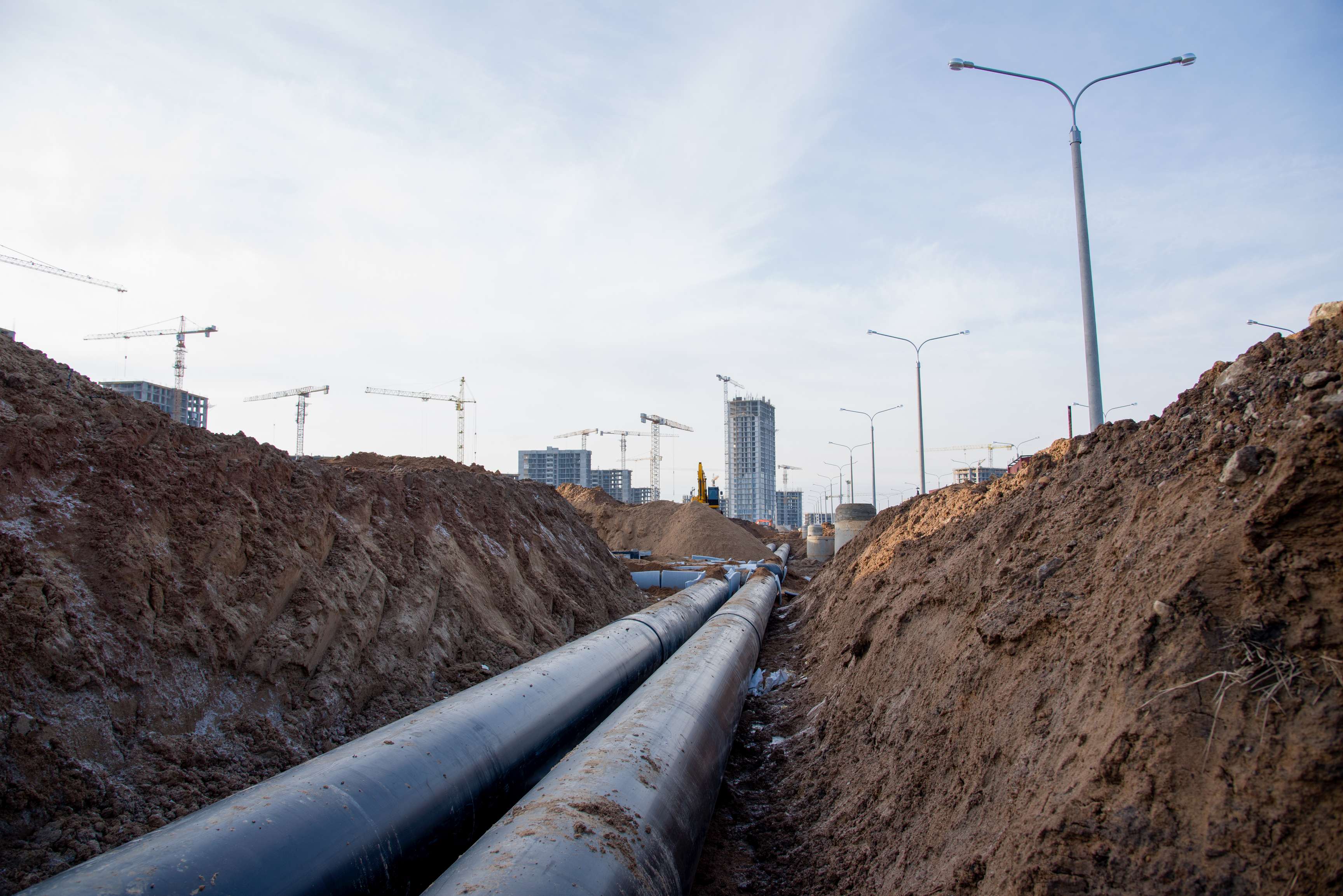 Two pipelines in the ground with backfilled earth on both sides, city with cranes in the background.