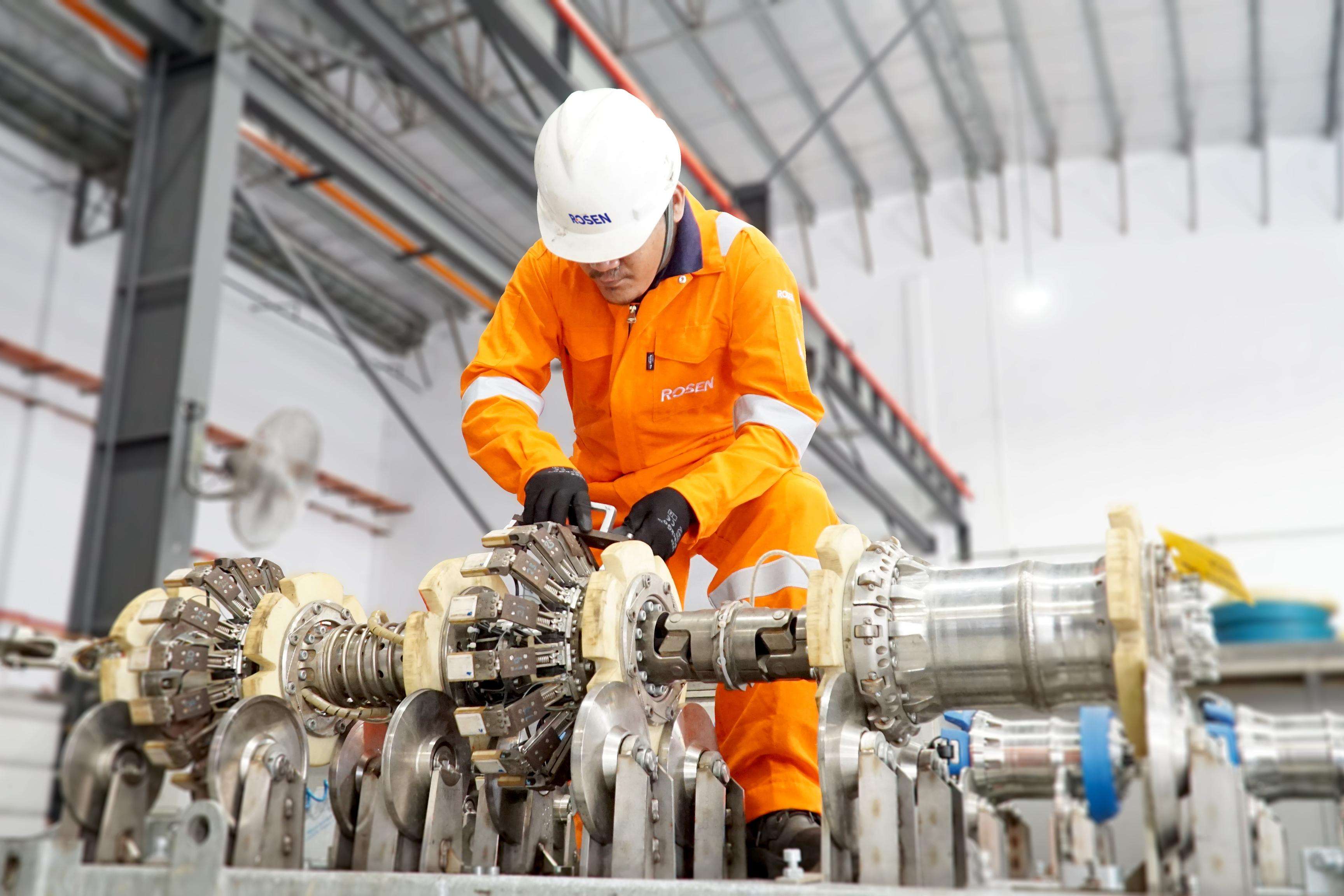 ROSEN employee with safety helmet and overall working on inspection equipment in a large factory hall.