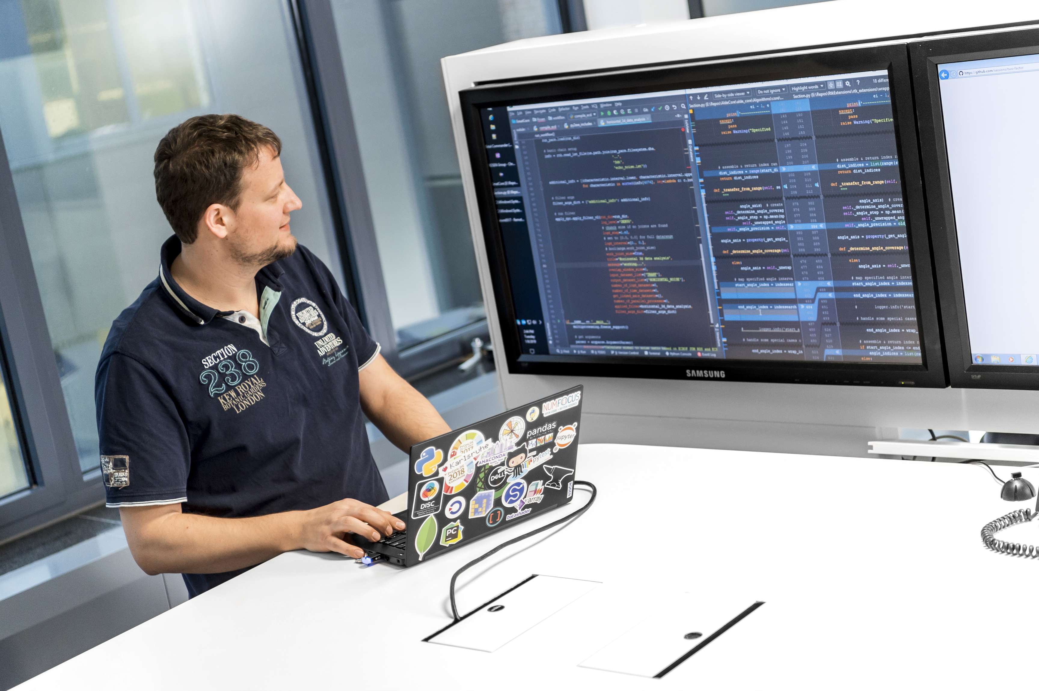 Ein Kollege aus der Abteilung für Softwareentwicklung steht an einem Stehtisch mit seinem Laptop und schaut auf den Präsentationsbildschirm.