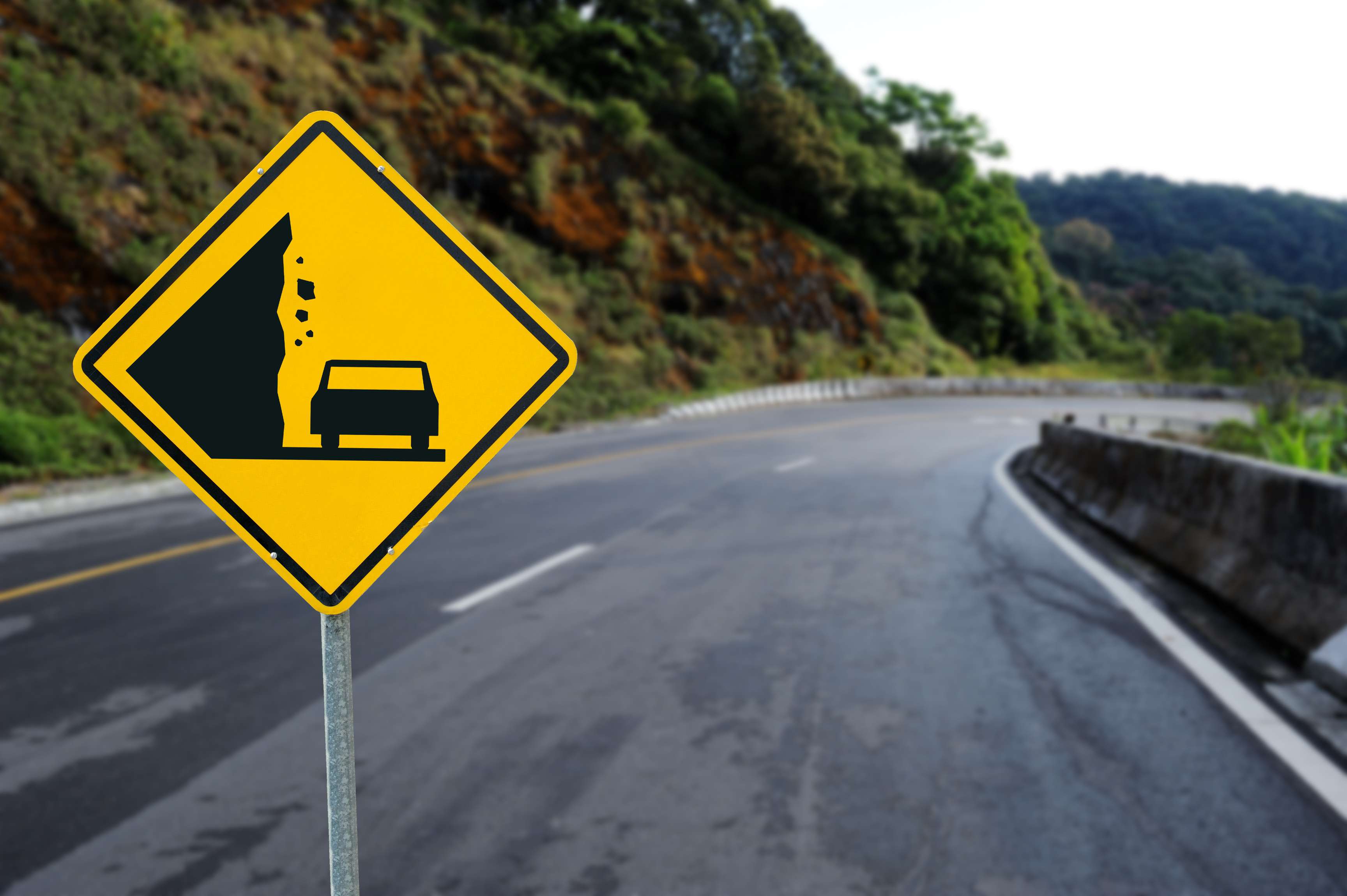 Yellow sign warning car driver of falling rocks on a road leading along a mountain.