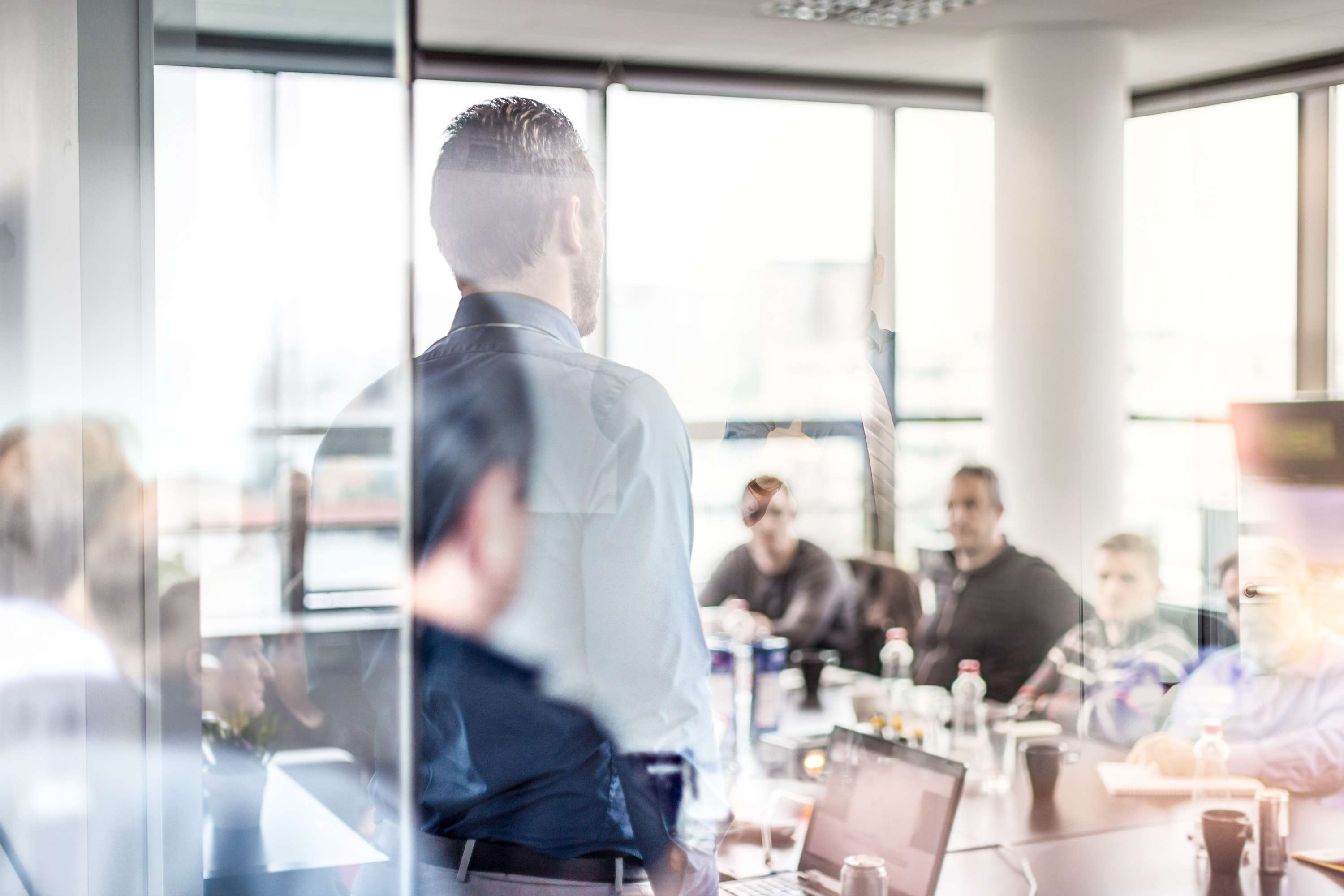 View through a pane of glass of a fully occupied conference room in which an employee is standing and explaining something to the others.