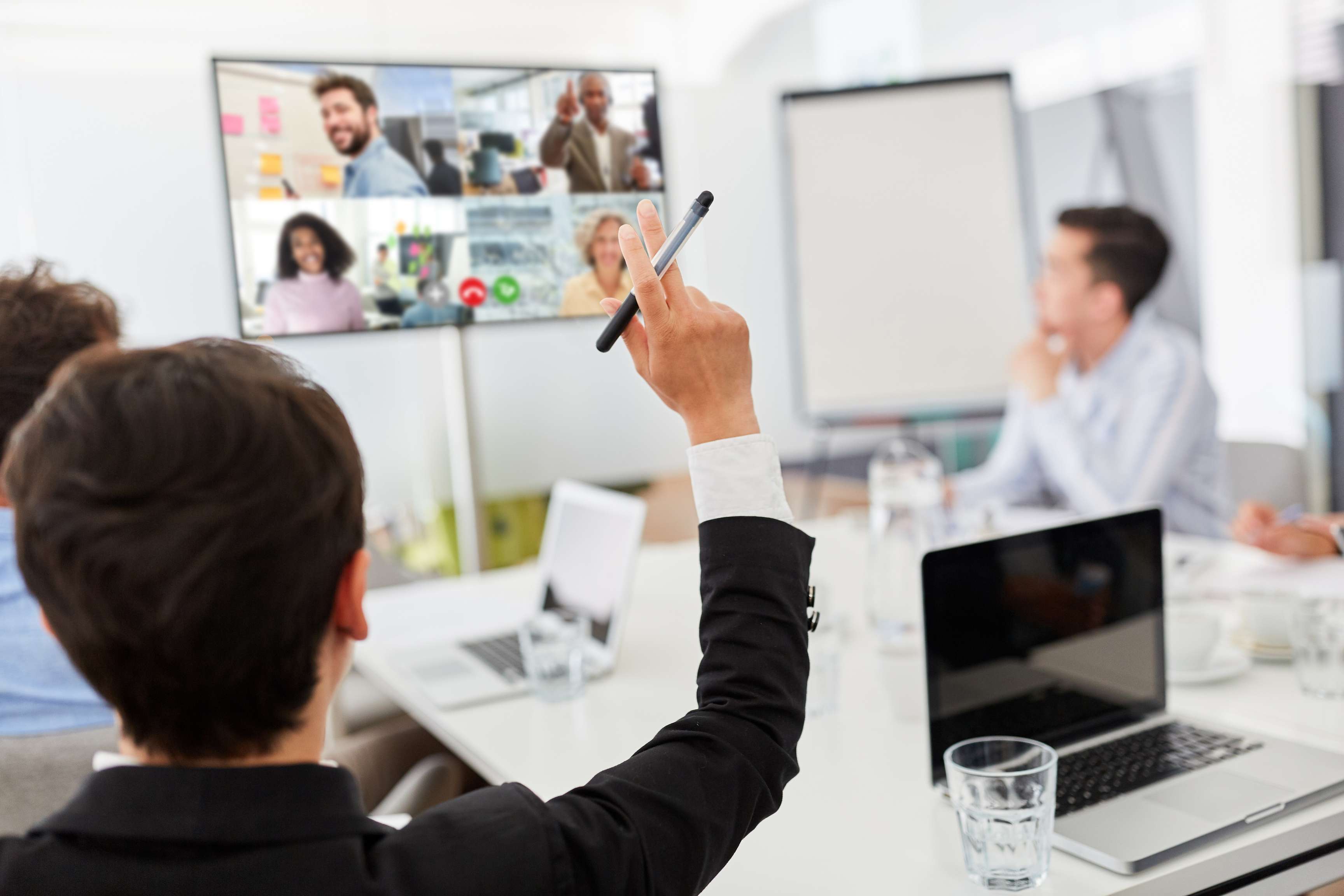 Several people in a meeting room looking at a screen showing people participating remotely. One person in the room raises his hand.