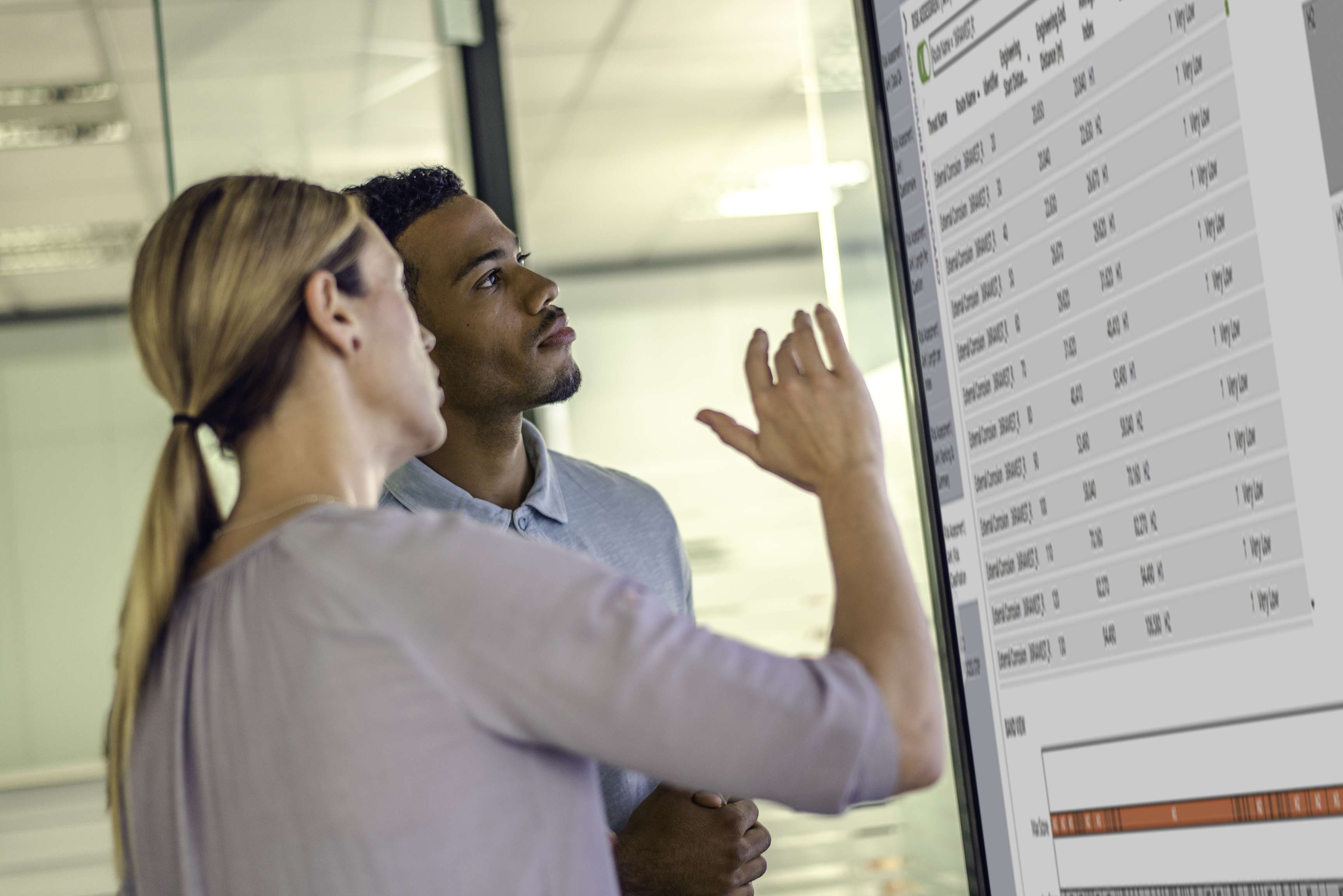 Two employees talking and pointing to a large screen with tables and data.