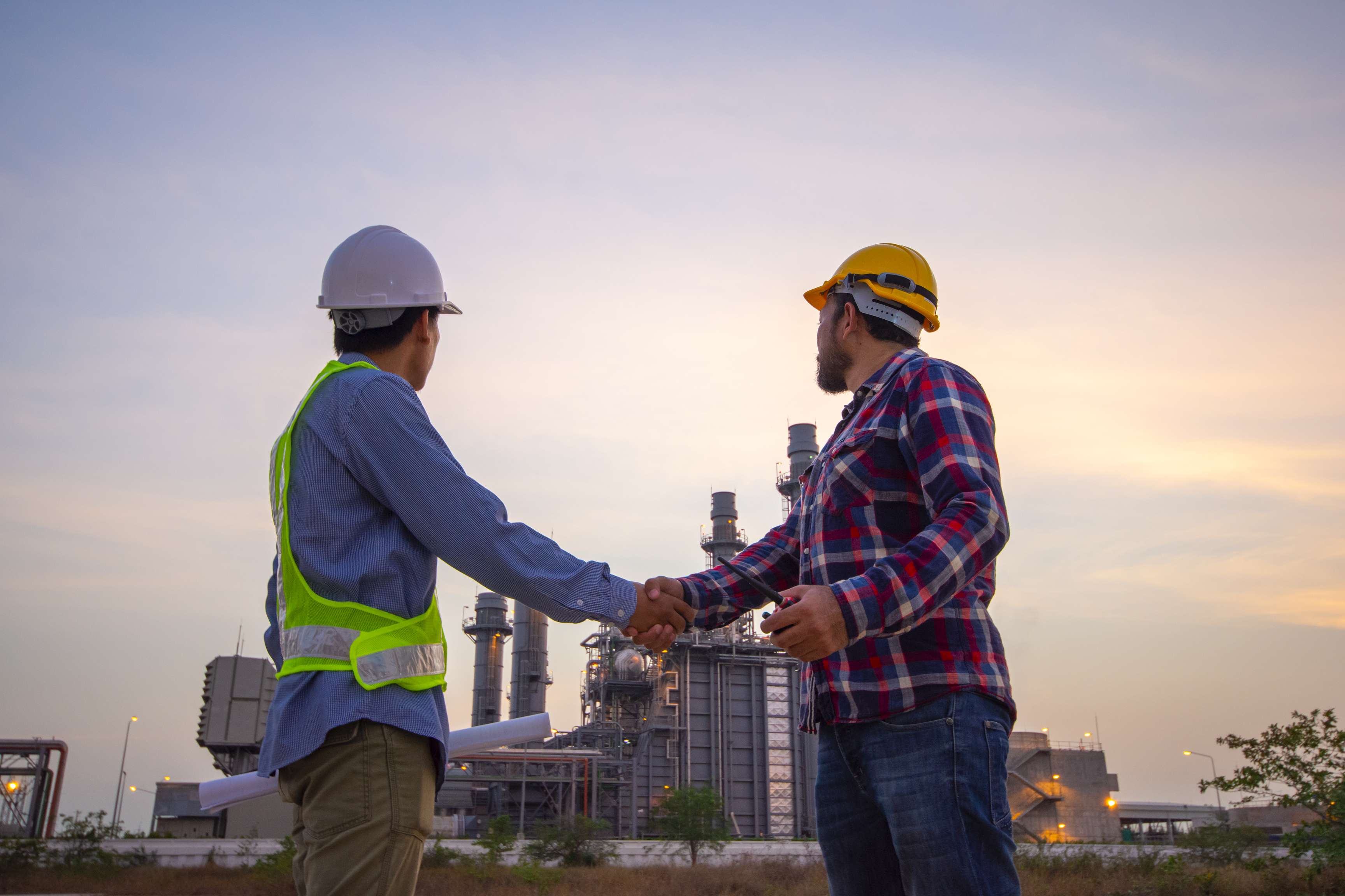 Two men in helmets shake hands in front of a refinery.