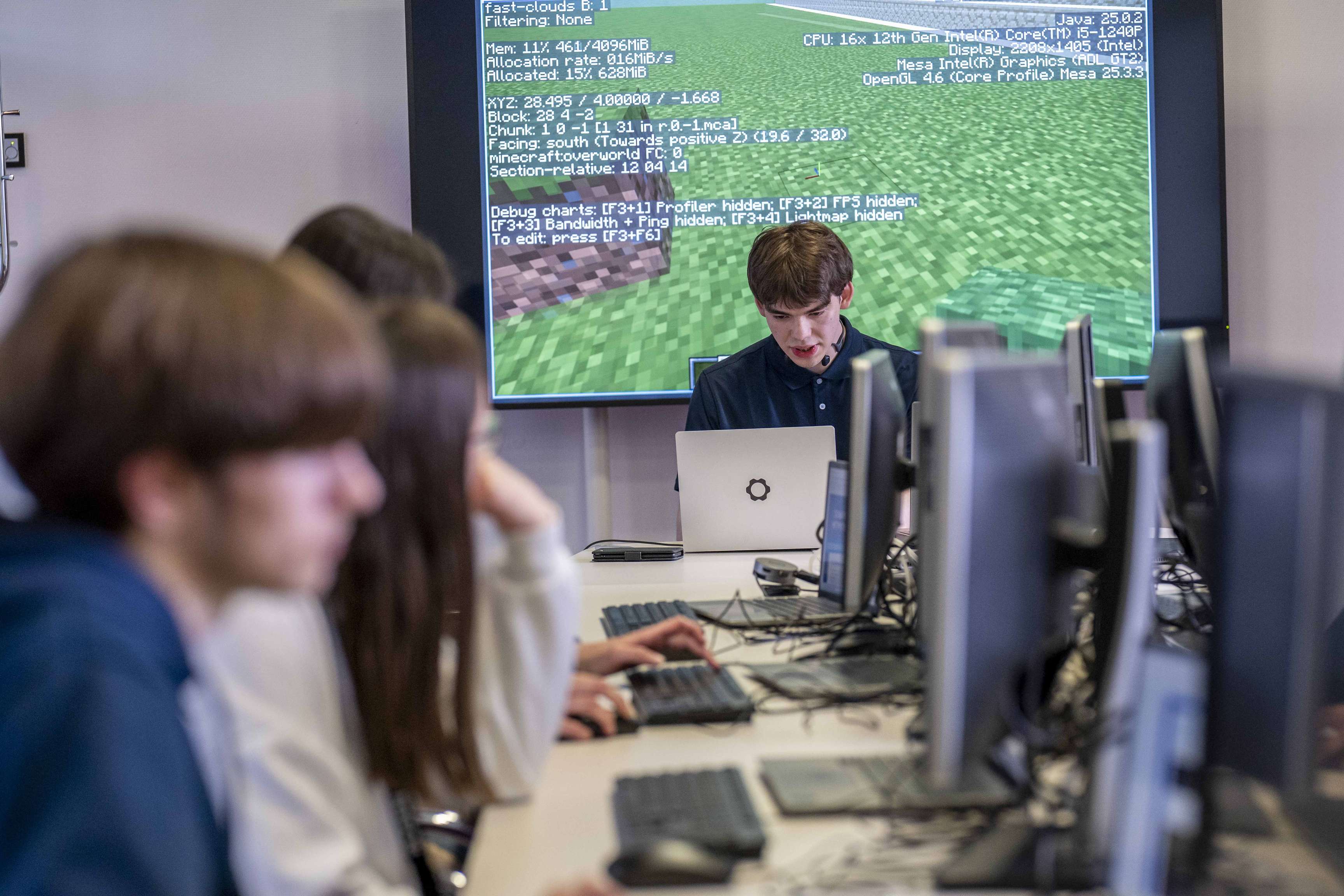 Schüler arbeiten an Computern im Klassenzimmer, während ein Referent auf einer großen Leinwand eine digitale Anwendung erklärt.