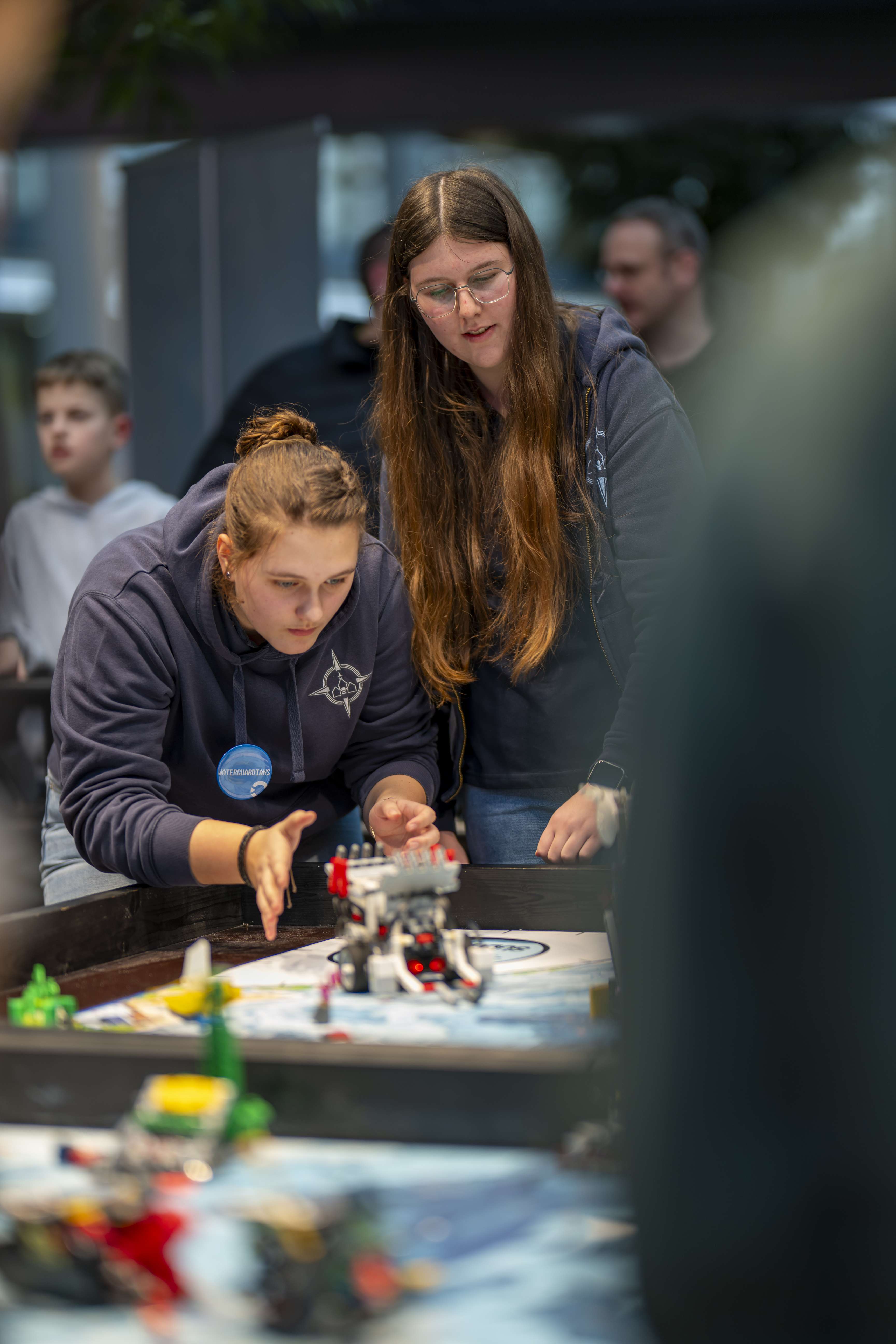 Two girls are standing in front of a table with a LEGO robot and observing its reaction.