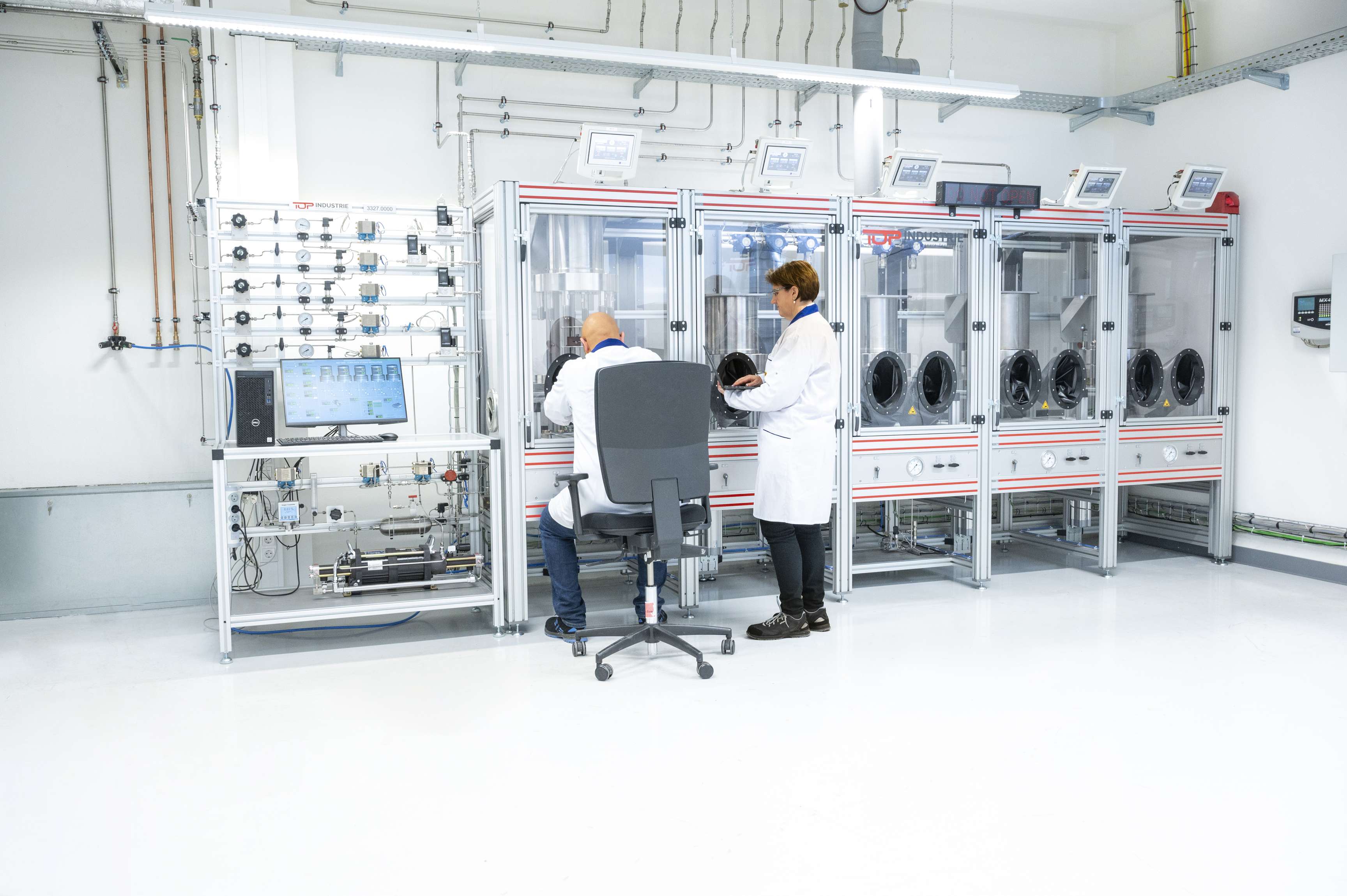 Image of a hydrogen testing lab with a man sitting and a woman standing with laptop in hand in front of the testing machines.