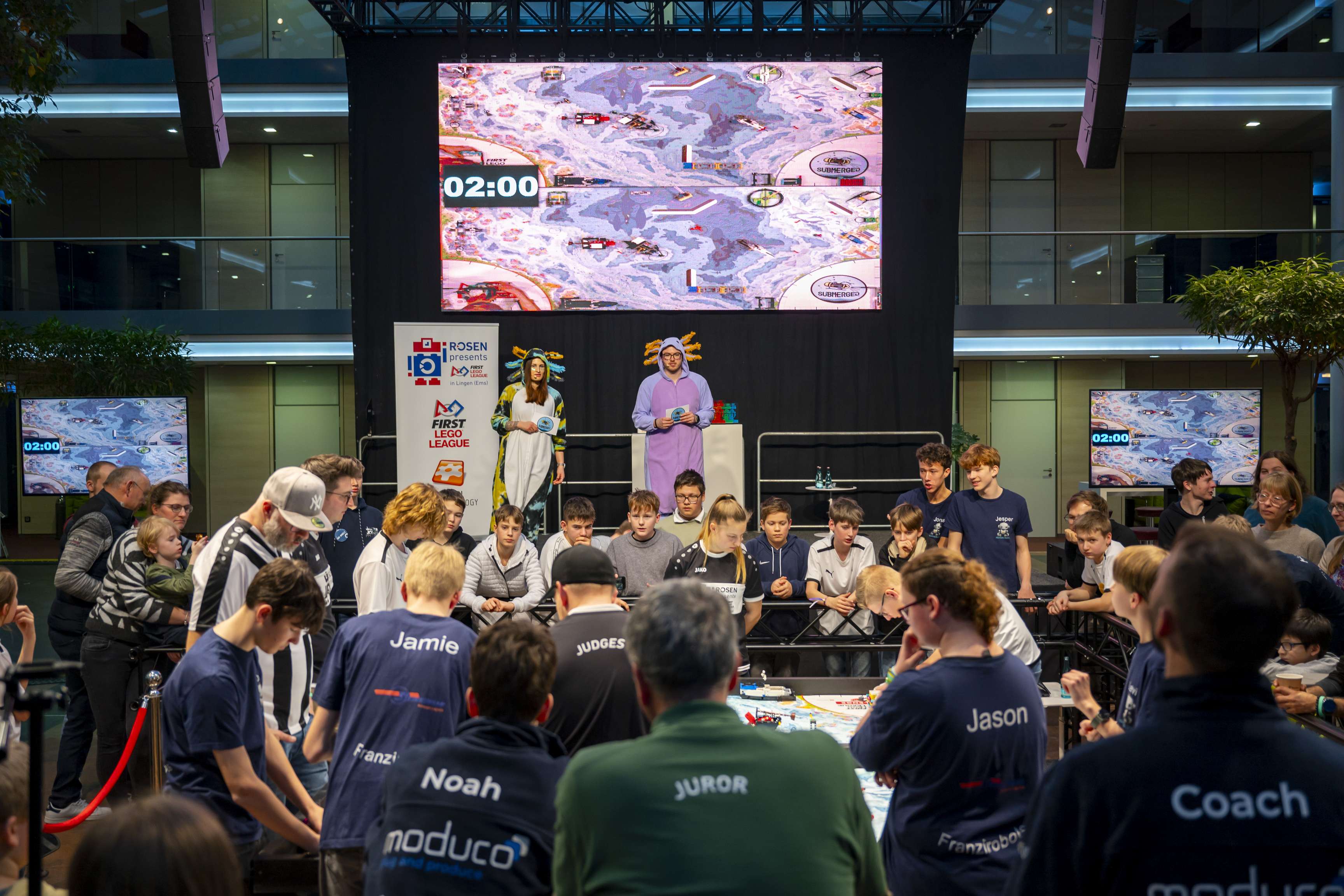 Two presenters in costumes are standing on the stage and watching the LEGO Challenge.