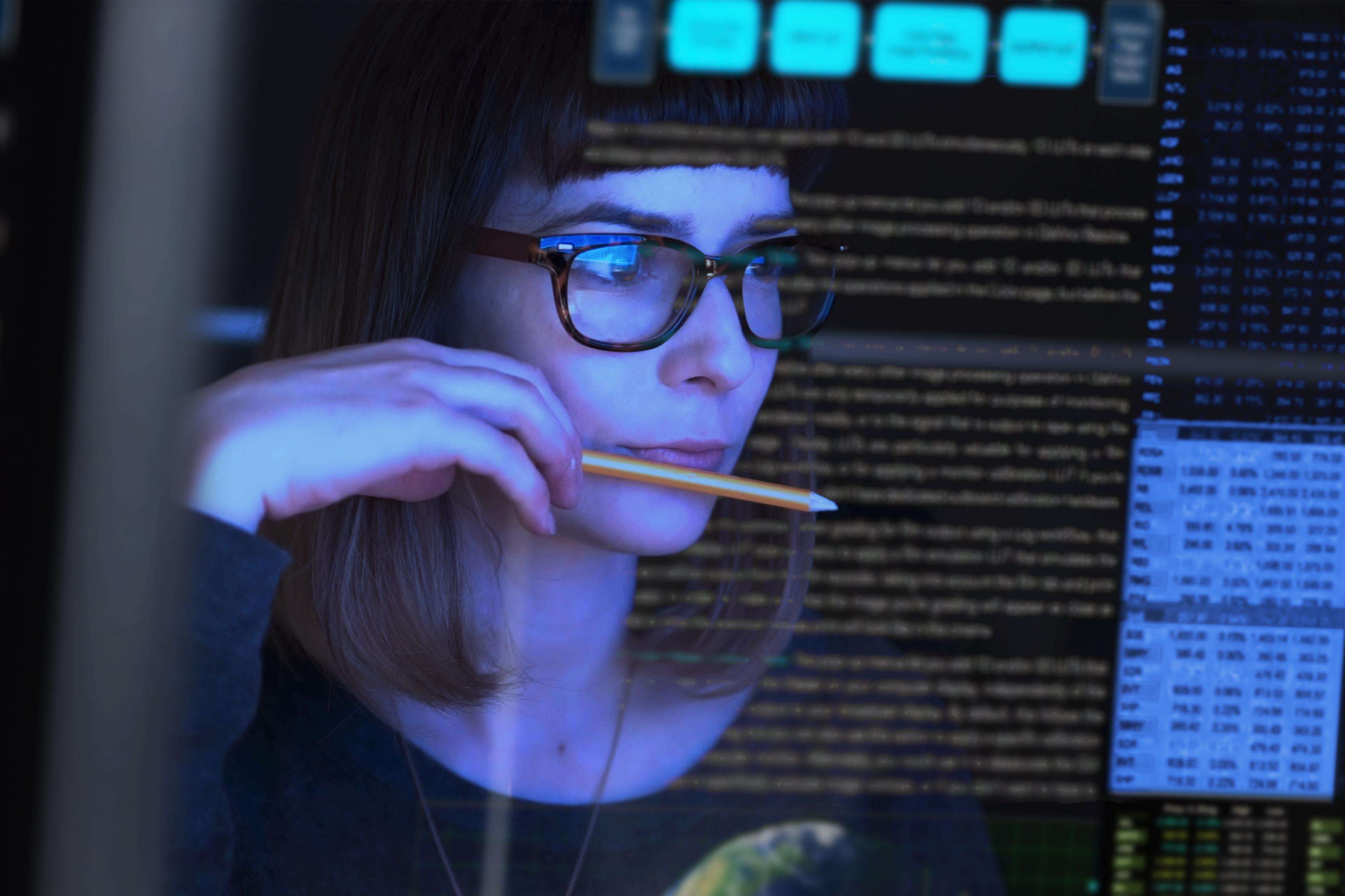Woman wearing glasses and holding a pencil in her hand and putting it to her lips while looking at a screen with data sets.