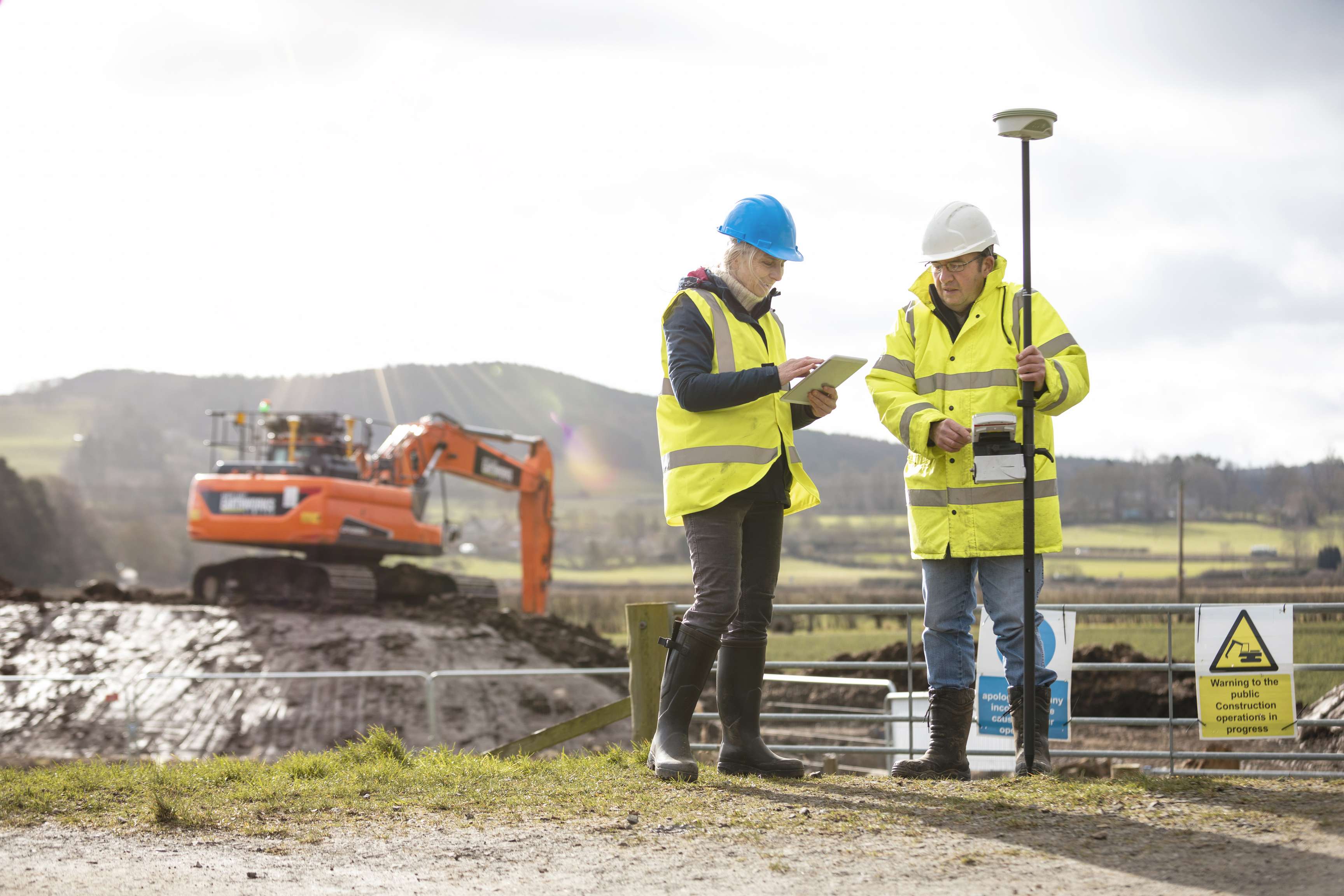 Two employees talk in front of a construction site with an excavator.