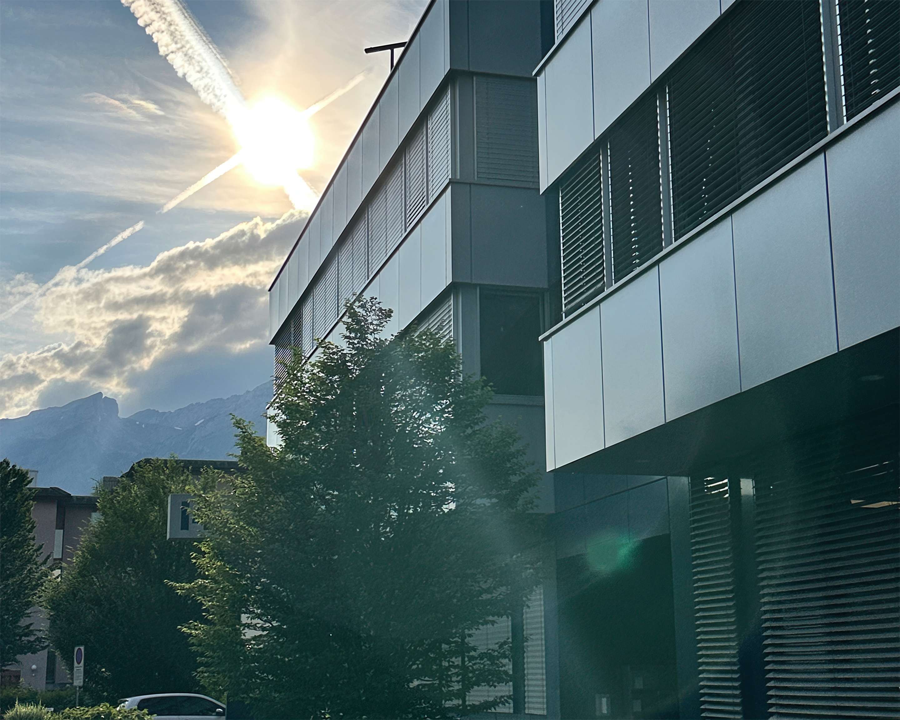 The photo shows the ROSEN Group headquarters in Switzerland, with its metal facade, backlit by the sun and surrounded by trees, with a mountain landscape in the background.
