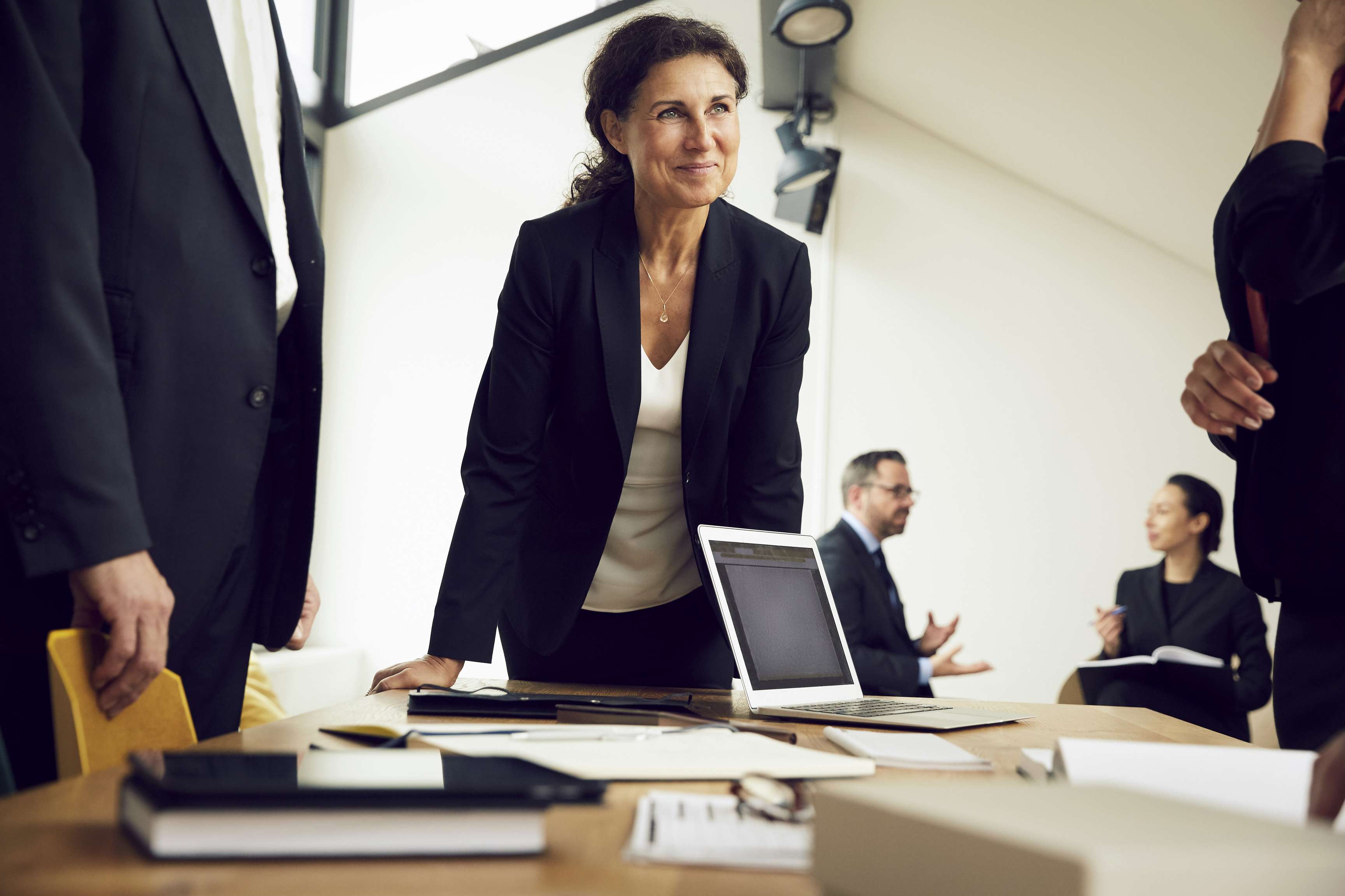 Colleagues stand in a group around a table with work materials, one colleague smiles friendly.