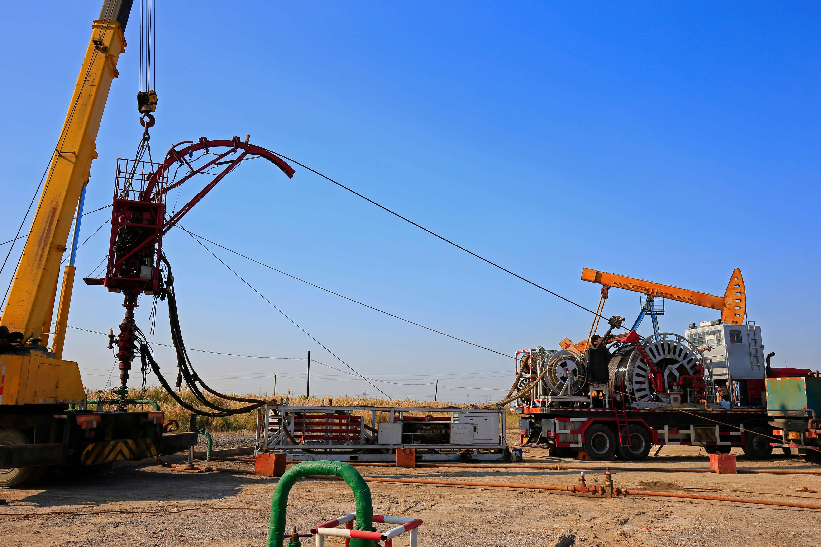 Coiled tubing operation with a crane on the left side and coiled tubing on the right side. Blue sky and oil pump in the background.