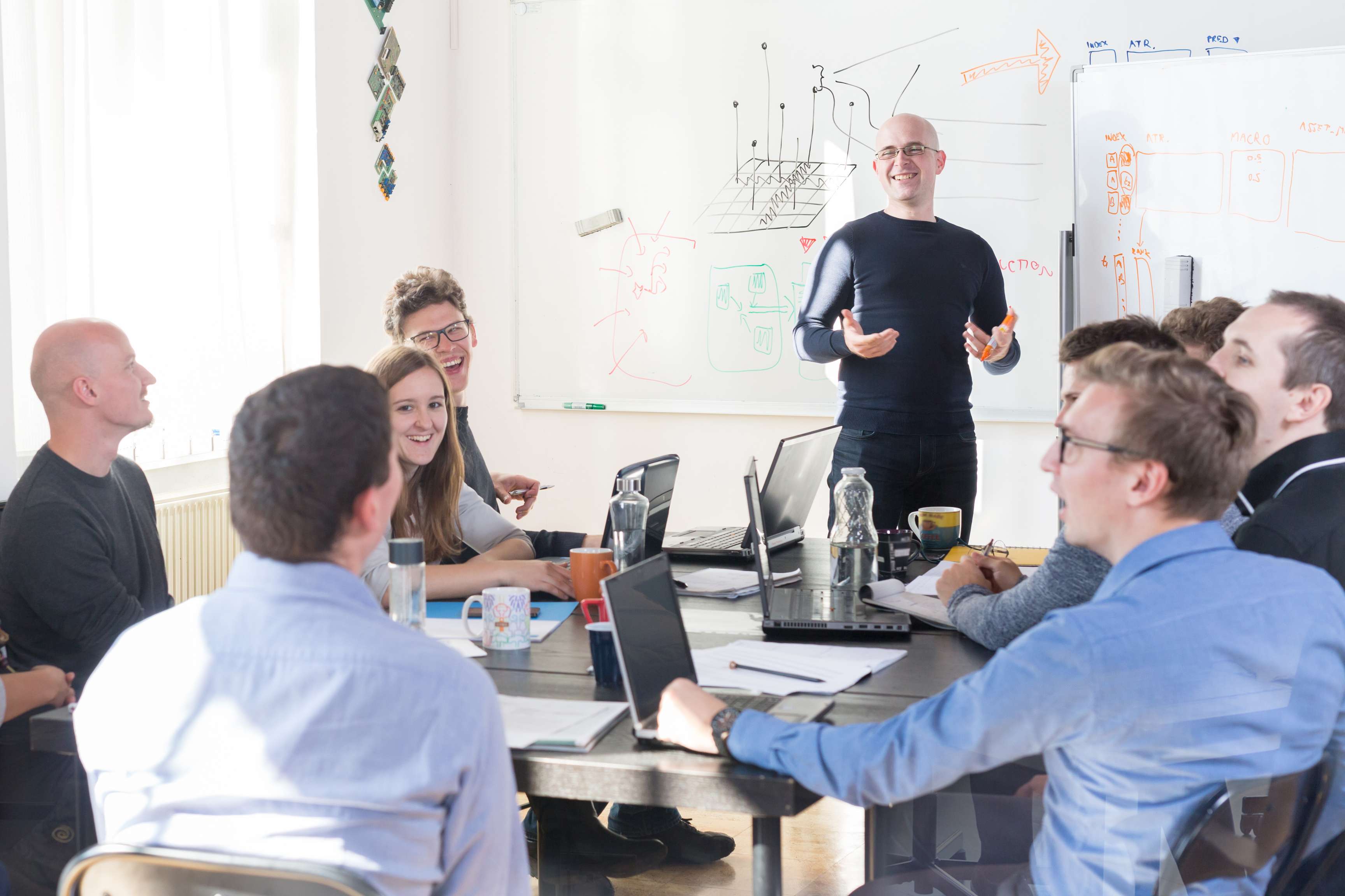 Many people sit in a meeting room around a table in front of their laptops while someone stands in front and presents something.