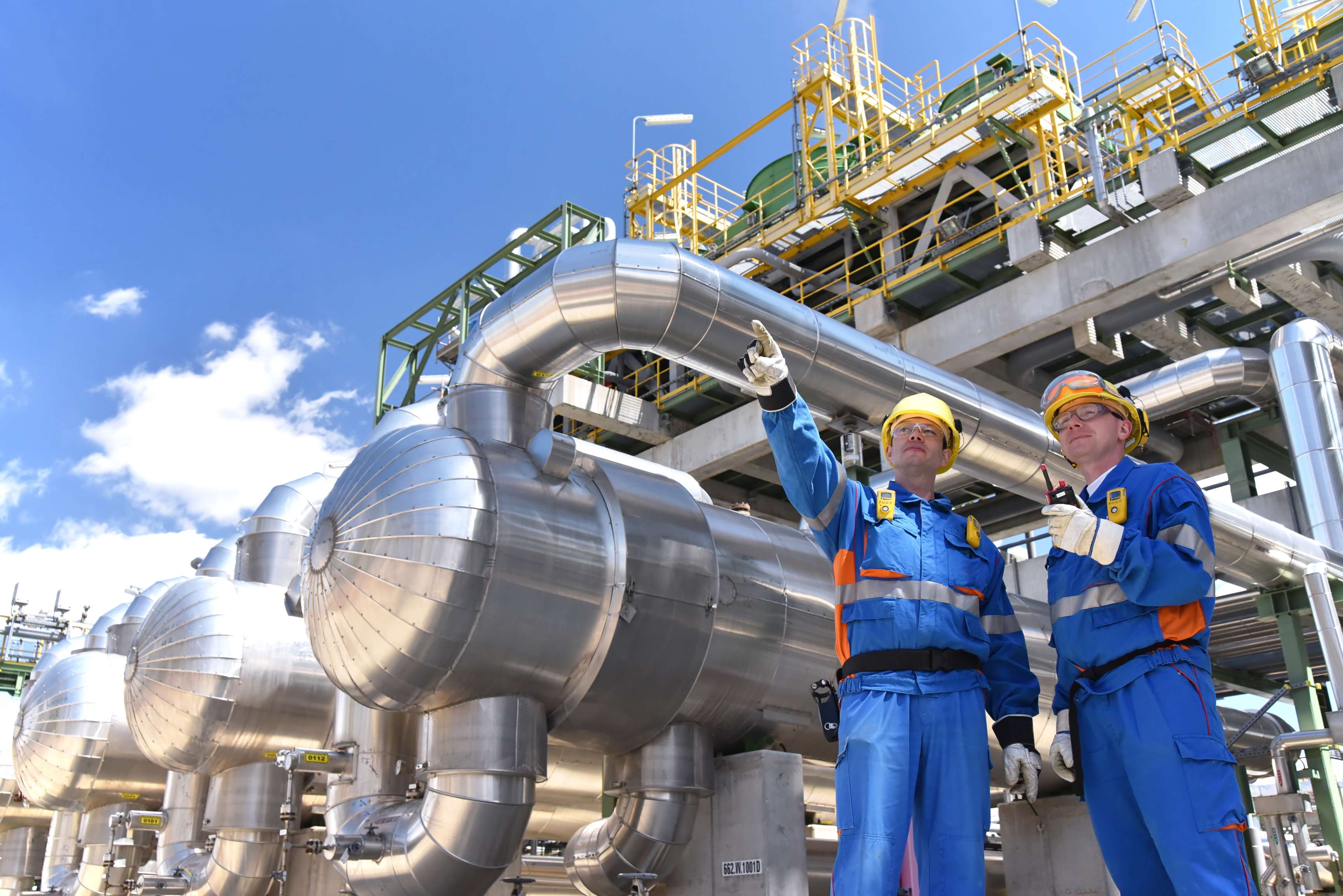 Two experts in front of a piping system and a pressure vessel, one of them pointing in one direction, the other holding a walkie-talkie.